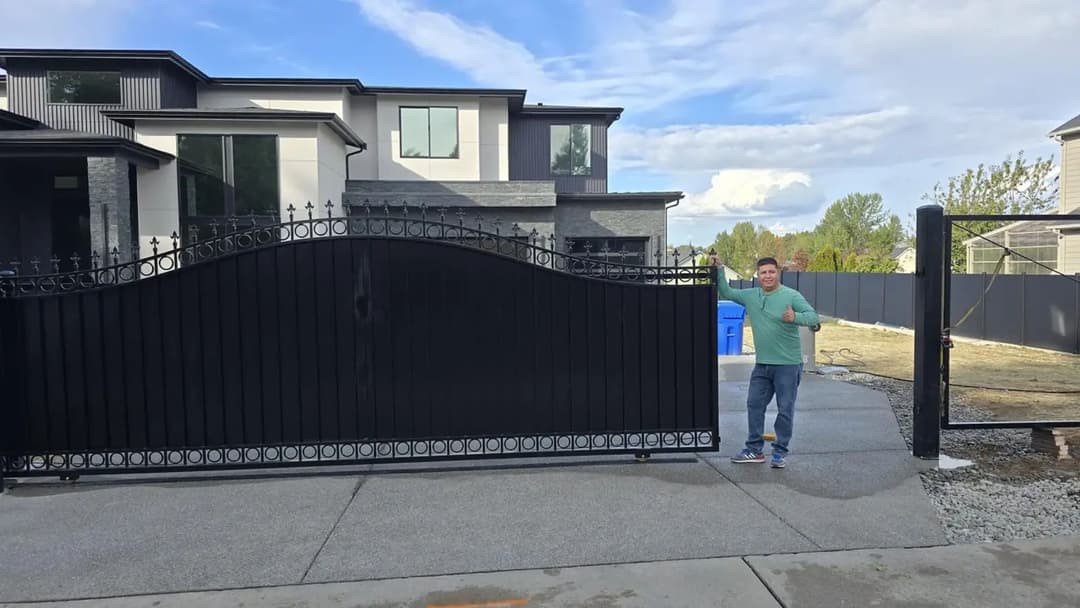 Man standing next to a decorative black gate of a modern home with blue recycling bins visible.