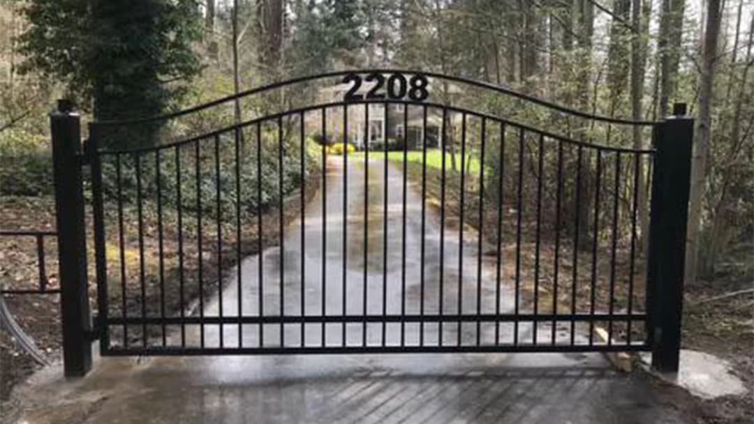 Black metal gate at entrance of driveway with house number 2208 surrounded by trees.