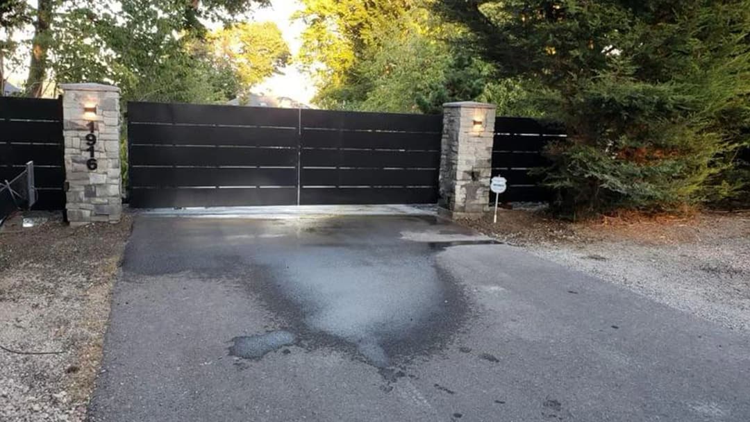 Black gated entrance with stone pillars, surrounded by greenery at a residential property.