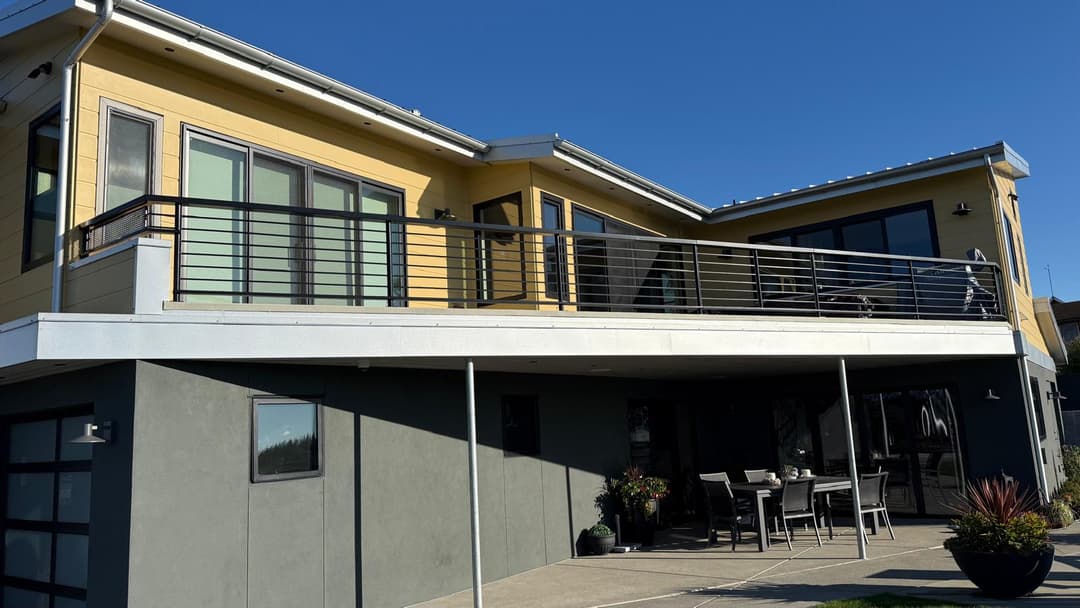 Modern yellow house with balcony, outdoor seating, and clear blue sky backdrop.