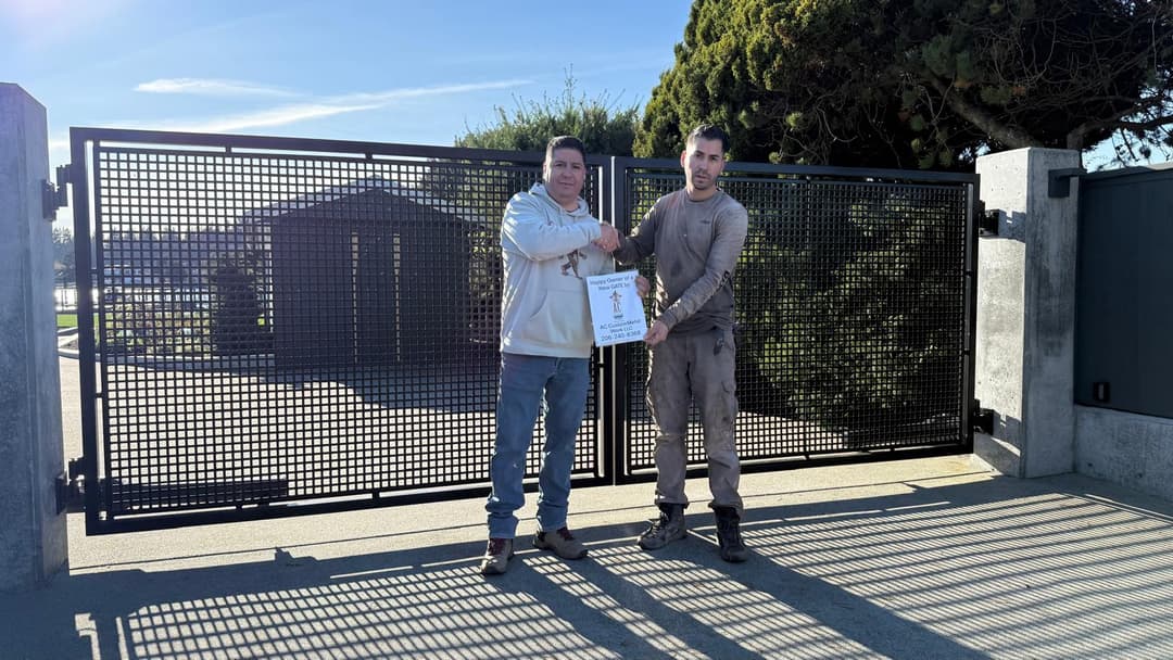 Two men shaking hands in front of a gated entrance, holding a certificate outdoors on a sunny day.