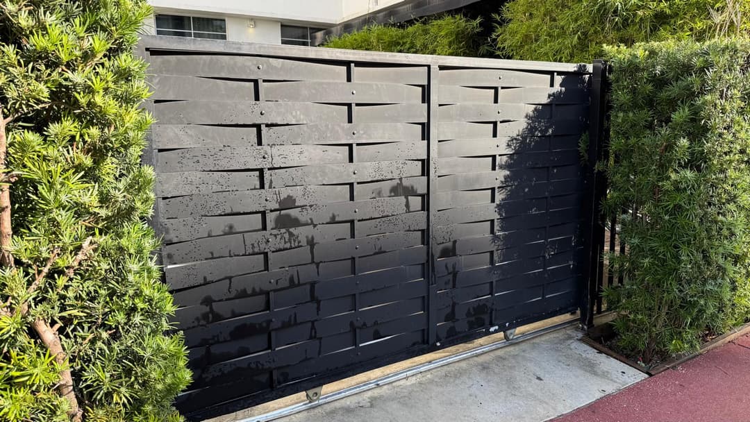 Modern black wooden gate surrounded by lush green shrubs in a residential area.