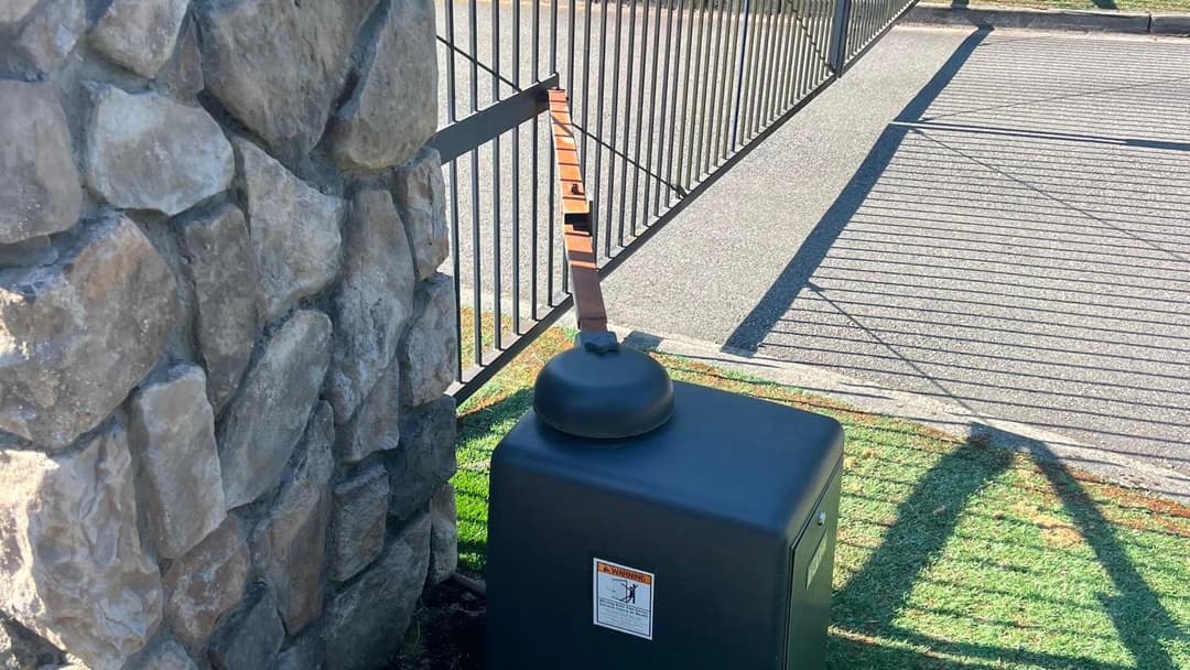 Black utility box beside a stone wall and gate, with a wooden strap attached.
