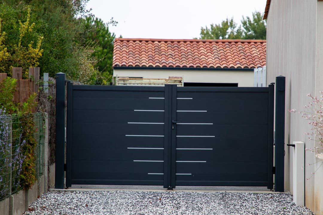 Modern black gate with horizontal lines, surrounded by greenery and a tiled roof home.
