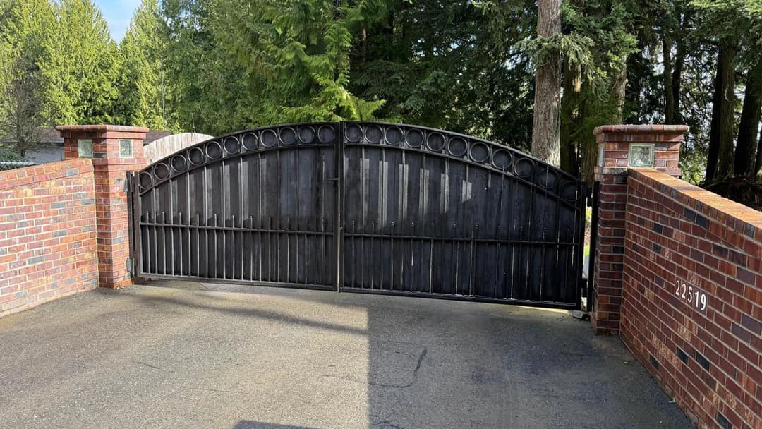 Black wrought iron gate with brick pillars, surrounded by lush green trees, leading to a driveway.