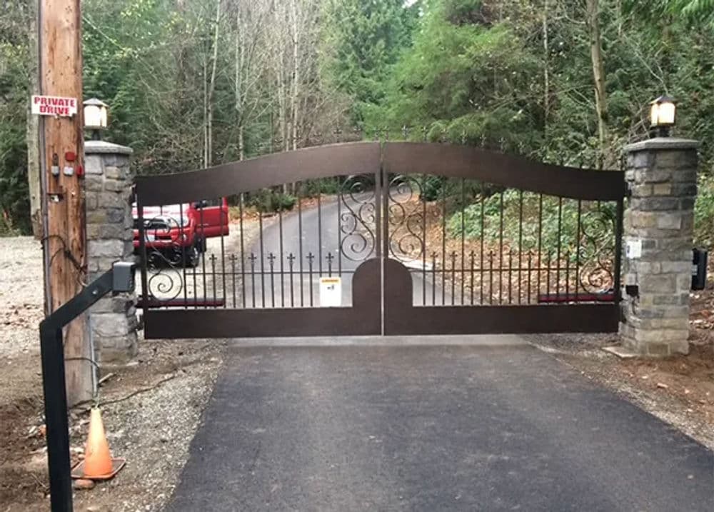 Iron access gate with stone pillars, leading to a private driveway surrounded by trees.